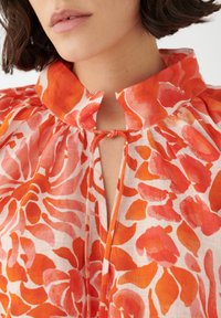 Blusa floreale in tessuto leggero con un motivo floreale rosso e rosa. Collo con apertura a chiave e un fiocco, dettagli plissettati sul colletto.