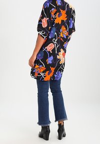Vloeiende zwarte bloemen tuniek met levendige oranje, paarse en blauwe patronen; gecombineerd met donkerblauwe uitlopende jeans en enkellaarsjes.