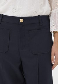 Blouse blanche en broderie florale rentrée dans un pantalon marine taille haute avec de grandes poches avant et un bouton doré, main reposant sur le côté.