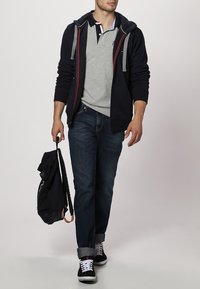 Hombre vestido con sudadera azul marino, polo gris, jeans oscuros y zapatillas negras, caminando mientras sostiene una mochila negra por una correa.