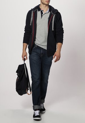 Man wearing navy hoodie, gray polo shirt, dark jeans, and black sneakers, walking and holding a black backpack by one strap.