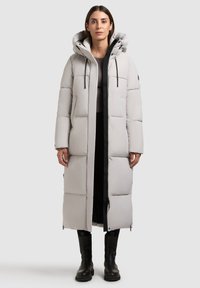 Light gray long puffer coat with a hood, featuring quilted sections, a front zipper, and black drawstrings, worn over a dark outfit.