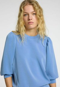 Gaiši zila blūze ar īsām, pūkainām piedurknēm un apaļu kakla izgriezumu. Gluda, maiga auduma ar smalku spīdumu un sānu šuvēm.