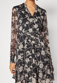 Svart klänning med vita blommönster och repmönster, genomskinliga långa ärmar, V-ringning och en knuten midjebälte, bärs av en person.