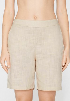 Pantalones cortos de lino beige hasta la rodilla, usados sobre el vientre descubierto, con bolsillos laterales y una cinturilla plana.
