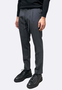 Pantaloni grigi su misura con una texture liscia, caratterizzati da pieghe frontali e una cintura elastica con coulisse. Abbinati a scarpe nere.