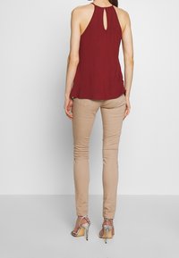 Bordowy top bez rękawów z dekoltem w kształcie łezki, zestawiony z beżowymi dopasowanymi spodniami i srebrnymi szpilkami, podkreślając casualowy look.