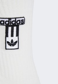 White ribbed sock featuring a black embroidered logo with three stripes and a trefoil at the base. Smooth texture, mid-calf length.