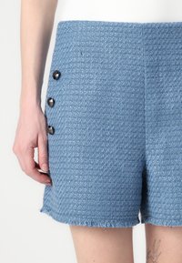 Pantaloni corti in tweed blu con una finitura testurizzata, caratterizzati da bordi sfilacciati e tre dettagli con bottoni neri sul lato anteriore.