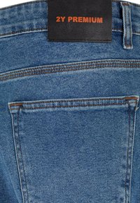 Denim jeans v svetlo modri barvi z teksturirano površino, z oranžnimi šivi in črno usnjeno oznako z napisom "2Y PREMIUM" na pasu.