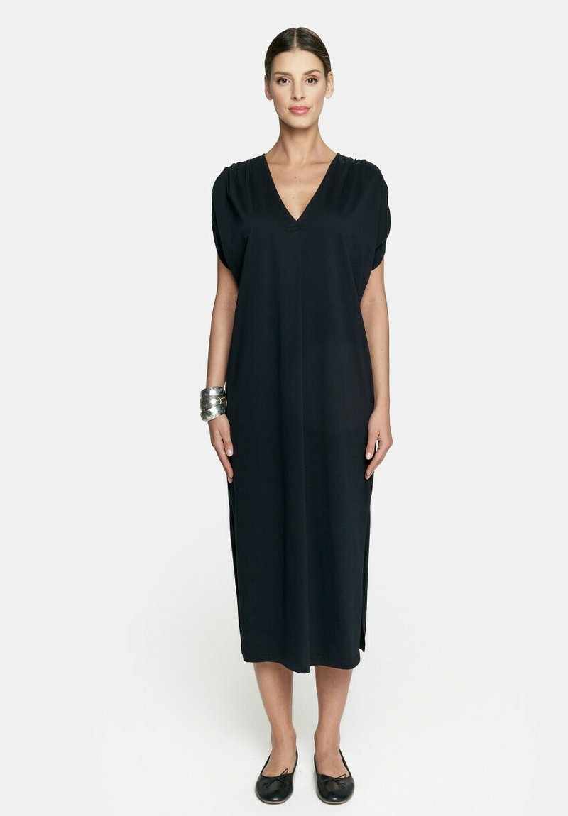 Czarna midi sukienka z dekoltem w kształcie litery V i krótkimi rękawami. Miękka tkanina z rozporkami po bokach. Płaskie czarne buty i srebrna bransoleta na nadgarstek.