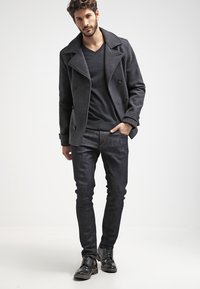 Palton peacoat gri închis, cu rabaturi mari și nasturi dubli, asortat cu un tricou negru cu decolteu în V și blugi închiși la culoare, completat cu pantofi negri.