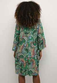 Zelena obleka s paisley vzorcem, s tremi četrtinskimi širokimi rokavi, plastičnim dizajnom ter mešanico barvitih vzorcev in tekstur. Dolžina obleke je nad kolenom.