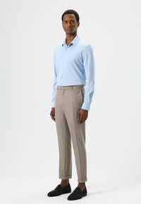 Hellblaues langärmliges Hemd kombiniert mit beigen, maßgeschneiderten Hosen mit Manschetten und schwarzen Loafers. Glatter Stoff mit klassischer Passform.