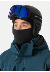 Skihjelm med sort finish, blåtonede goggles og en sort fleece halsvarmer. Personen har en teal jakke med høj krave på.