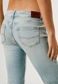 Jasnoniebieskie jeansy z denimu noszone przez osobę, pokazujące tylne kieszenie, detale przeszyć oraz brązową skórzaną naszywkę marki na pasku od spodni.