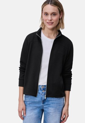 Kvinde iført en sort lynlåstrøje over en hvid skjorte og lyseblå jeans, stående mod en ensartet lys baggrund.