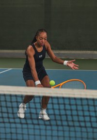 A tennis player in a dark blue sleeveless top and matching shorts is reaching for a green ball with an orange racket on a blue court.