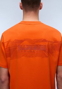 Orange kortærmet t-shirt med en tekstureret grafisk design og "NAPAPIRIRI" logo samt vandrette linjer på ryggen. Glat stof.