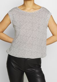 Femme portant un haut sans manches blanc avec des taches noires, rentré dans un pantalon en cuir noir, les mains reposant sur les hanches.