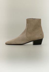 Μπεζ μπότες αστραγάλου από σουέντ με μυτερή μύτη, χαμηλό μαύρο τακούνι σε μπλοκ, πλευρικό φερμουάρ και μικρό χρυσό λογότυπο κοντά στη φτέρνα.