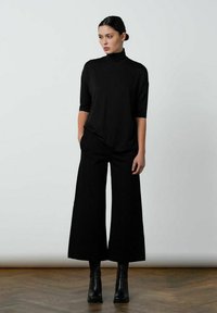 Zwarte coltrui met driekwartmouwen, gecombineerd met wijde zwarte broek en gestapelde zwarte enkellaarsjes op een gestructureerde houten vloer.