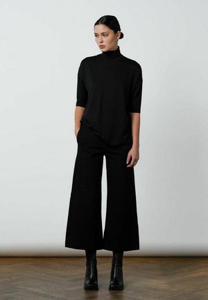 Sort turtleneck med trekvartærmer parret med brede sorte bukser og høje sorte ankelstøvler på et tekstureret trægulv.