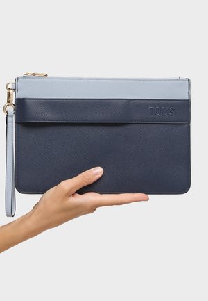 Mano sosteniendo un bolso de mano rectangular bicolor azul de cuero con cremallera y logo TOUS grabado al frente.