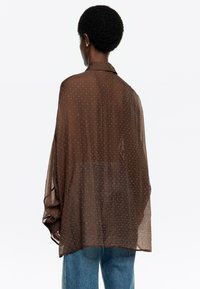 Camisa marrón fluida y transparente con un patrón de puntos, que cuenta con un cuello tipo solapa y mangas sueltas, combinada con pantalones de mezclilla azul claro.
