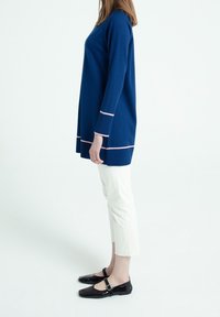 Maglione tunica di maglia blu navy con dettagli rosa pallido sui polsini e sull'orlo, abbinato a pantaloni bianchi a 3/4 e scarpe Mary Jane nere in vernice.