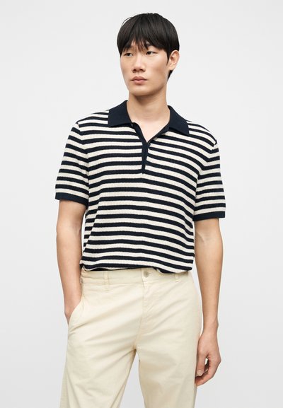 Polo à manches courtes rayé bleu marine et crème, avec col plat et tissu texturé. Associé à un pantalon beige clair.
