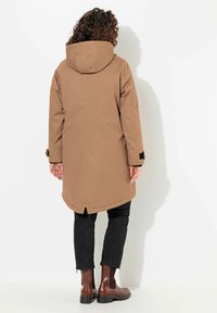 Manteau imperméable marron avec capuche, manches longues et ourlet incurvé. Pantalon noir à motifs et bottines marron.