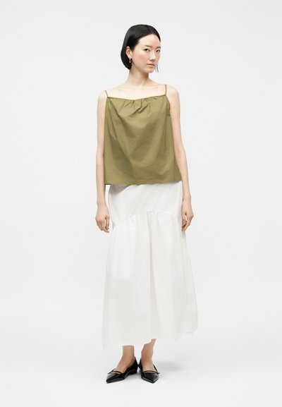 Γυναίκα με ελιάς πράσινο, αμάνικο τοπ, λευκή ροή φούστα μέχρι τους αστραγάλους και μαύρα μυτερά φλατ παπούτσια, στέκεται μπροστά σε απλό φωτεινό φόντο.