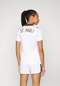 Hvit sportsdrakt med brun "ST. PAULI" tekst på baksiden. Korte ermer og rund krage, sammen med hvite shorts. Glatt stoffstruktur.