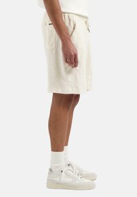 Shorts bej din material texturat, cu un model matlasat, având buzunare laterale și talie cu șnur. Purtați cu șosete albe și adidași.