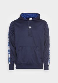 Laivastonsininen Nike-huppari kengurutaskulla, säädettävällä huppuosalla ja valkoisella Nike-logolla rinnassa sekä kuvioiduilla raidoilla hihoissa.