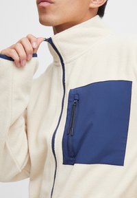 Beige Fleecejacke mit hohem Kragen, ausgestattet mit einer dunkelblauen, mit Zipper versehenen Brusttasche und marineblauen Akzenten an Reißverschluss und Ärmeln.