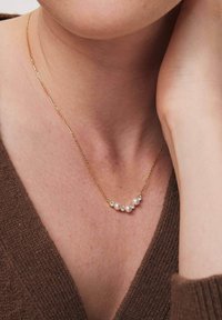 Złoty naszyjnik łańcuszkowy z pięcioma małymi perłami i błyszczącymi akcentami, spoczywający na skórze, w tle z teksturowanym brązowym topem.