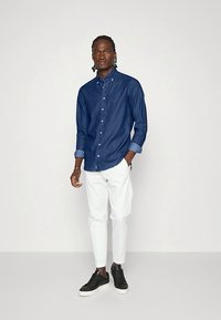 Vyras su pintomis plaukų sruogomis dėvi tamsiai mėlyną marškinių tipo palaidinę, baltas kelnes ir juodus sportinius batus, stovintis prieš paprastą šviesią fono.