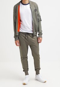 Tuta verde oliva con interni arancioni, composta da una giacca con zip e pantaloni con cordino. Scarpe da ginnastica bianche completano l'outfit.