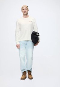 Camisa de manga larga crema con jeans azul claro, botas marrones y un gorro beige; sosteniendo un casco negro.