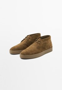 Bottines chukka en suede marron avec un bout rond, un design à lacets et une surface texturée. Équipées d'une semelle en caoutchouc contrastante pour une plus grande durabilité.