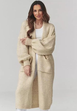 Kobieta ubrana w długi beżowy, dziergany kardigan z dużymi rękawami i przednimi kieszeniami, stojąca na tle jasnoszarego tła.