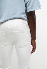 Witte denim jeans met achterzakken, hoge tailleband, subtiele stiksel en een stoffen label aan de achterkant. Lichtblauw T-shirt gedeeltelijk zichtbaar.
