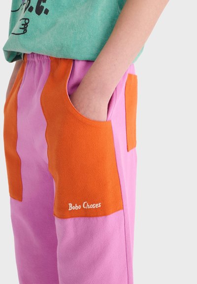 Personne portant un pantalon rose avec de grandes poches orange, une main à l'intérieur de la poche, chemise verte partiellement visible au-dessus.