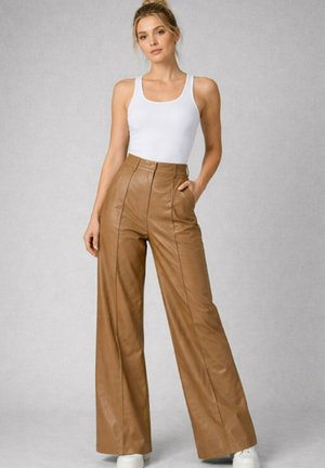 Femme debout portant un haut blanc sans manches et un pantalon large taille haute en cuir marron, les mains dans les poches.