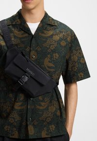 Sort sling taske med lynlåslukning og "HUGO" logo, båret over en mønstret mørkegrøn skjorte med fugle og blomster motiver.