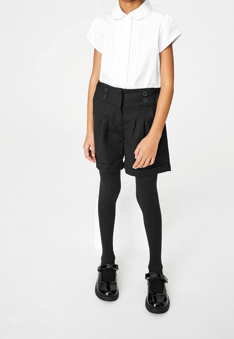 Enfant portant une chemise blanche à manches courtes, un short taille haute noir avec des boutons, des collants noirs et des chaussures Mary Jane noires brillantes se tenant sur un fond blanc.