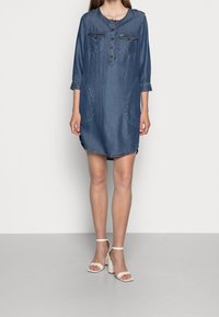 Denimmekko, jossa on pyöreä kaula-aukko, kolme neljänneksen hihat, kaksi rintataskua ja kaarevat sivusaumat. Tässä mekossa on näkyvät saumat ja rento istuvuus.