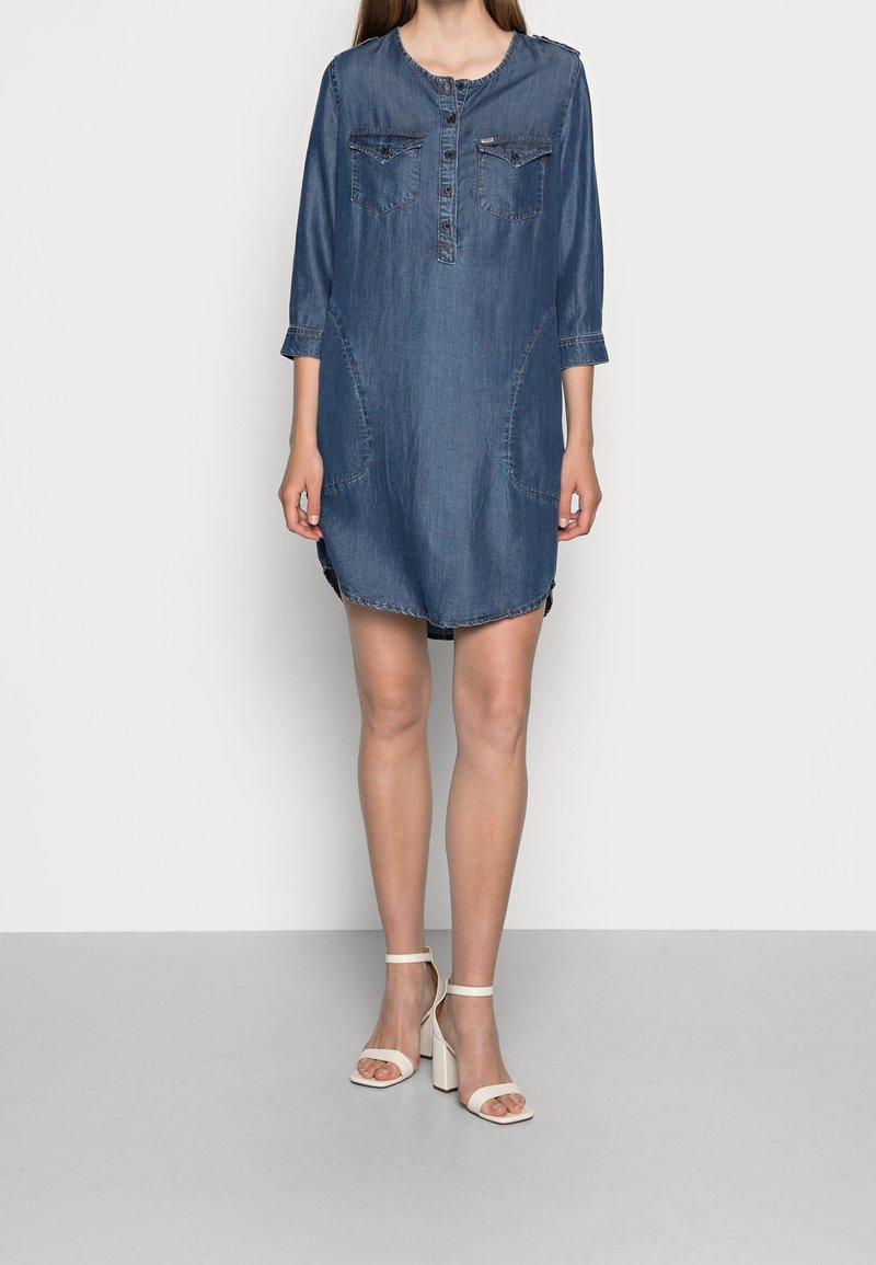 Denimmekko, jossa on pyöreä kaula-aukko, kolme neljänneksen hihat, kaksi rintataskua ja kaarevat sivusaumat. Tässä mekossa on näkyvät saumat ja rento istuvuus.